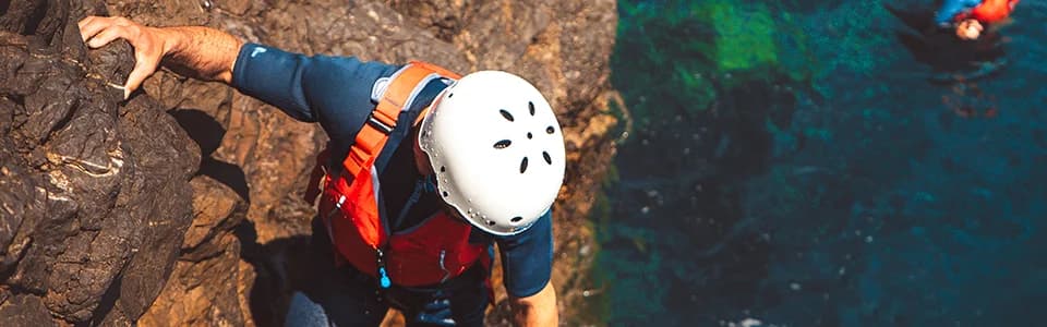 Coasteering participant wearing helmet and safety gear descending coastal rocks above deep blue water in Ibiza